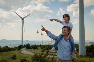 爸爸带着女儿坐在肩膀上看着风力涡轮机，微笑着。